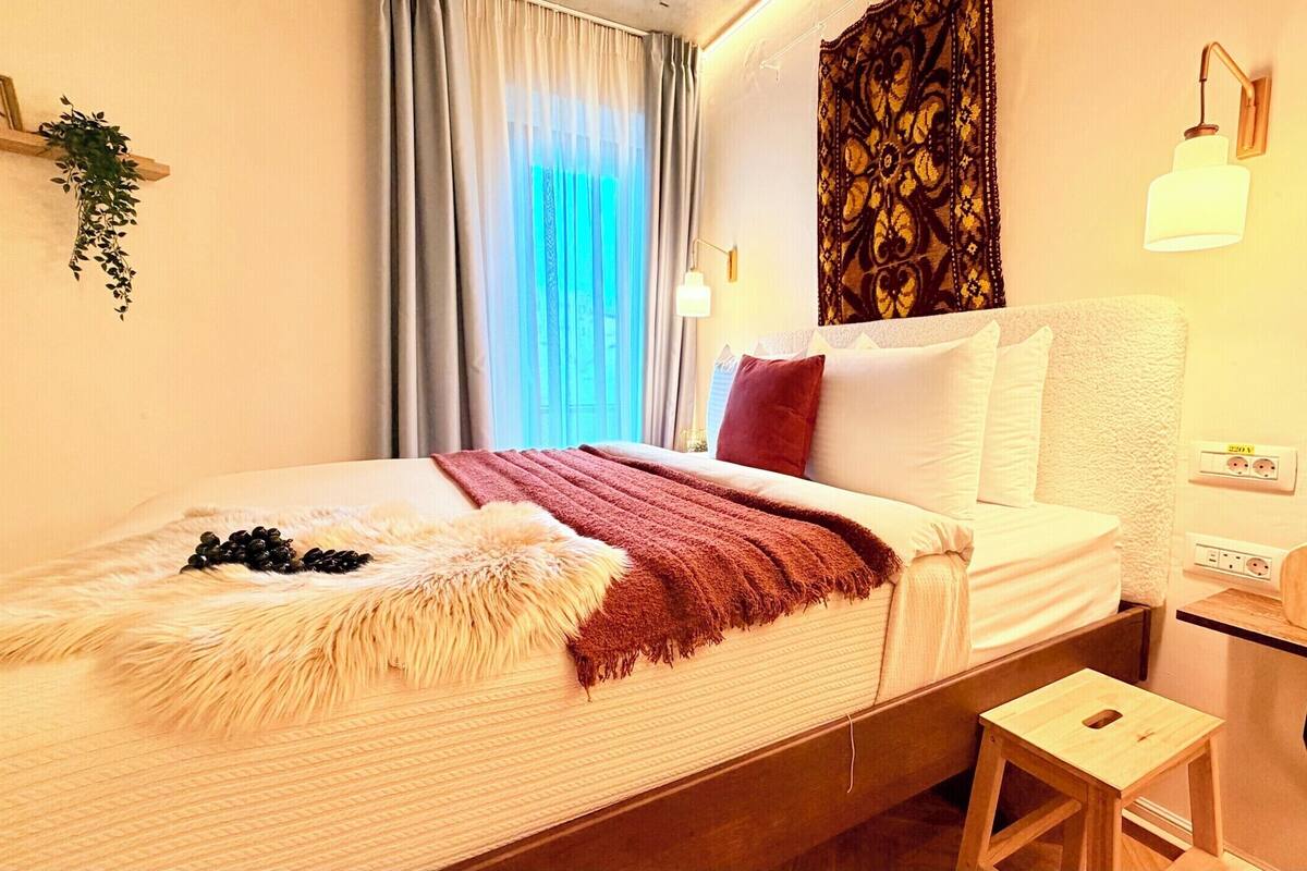 A hotel room with a bed, a nightstand, a wall-mounted lamp, a power outlet, and a window with curtains.