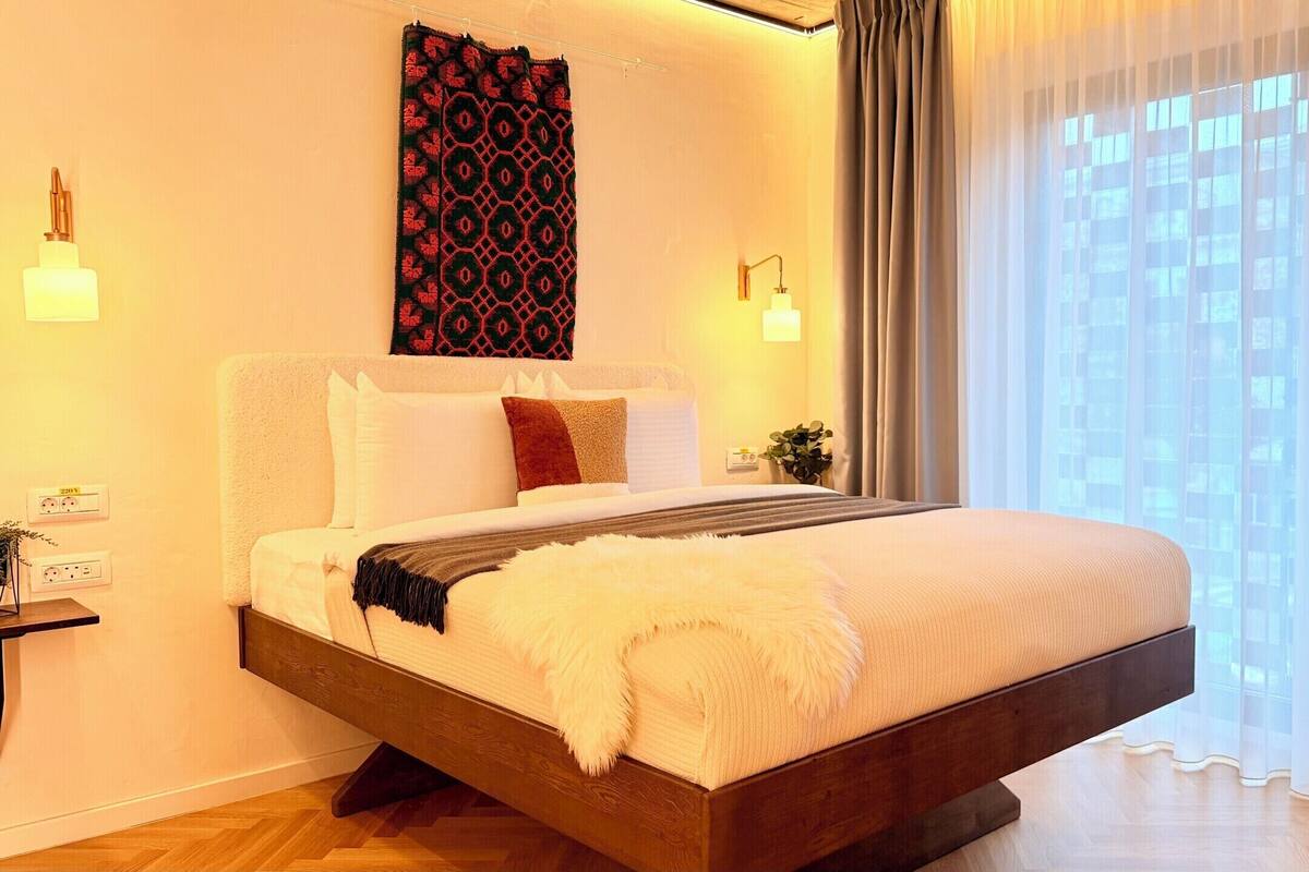 A modern hotel room with a bed, bedside lamps, a view of the city, and a window with sheer curtains.