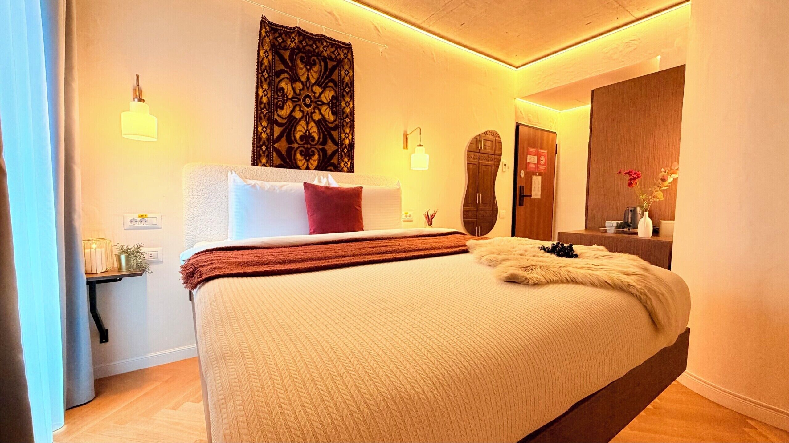A hotel room with a large bed, a nightstand, a wall-mounted lamp, a tapestry, and a mirror.