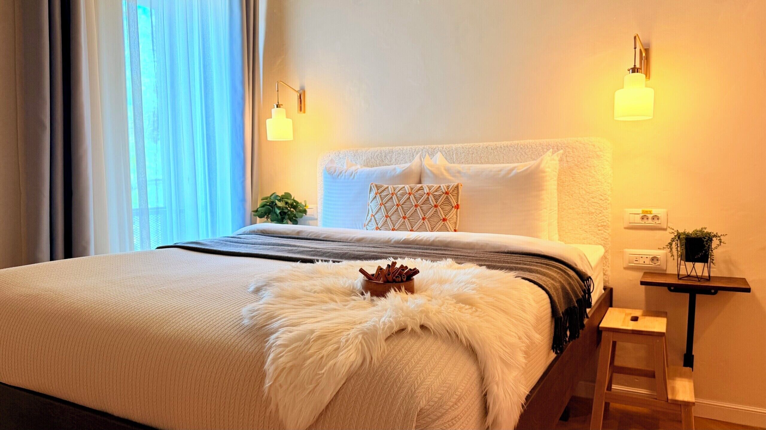 A neatly made bed with a fluffy white throw, a patterned pillow, and a small plant on a bedside table.