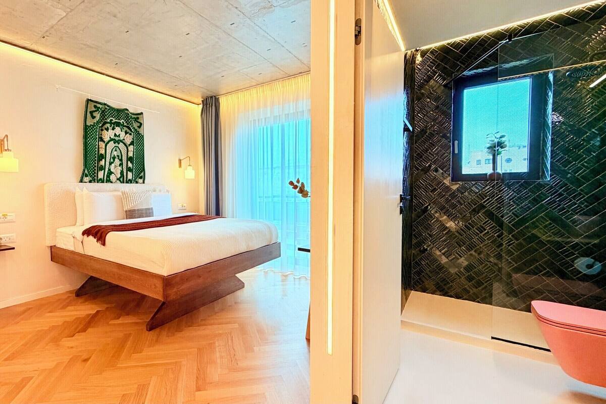 A modern bedroom with a wooden bed, a green tapestry, a wall-mounted light, and a large window with curtains.