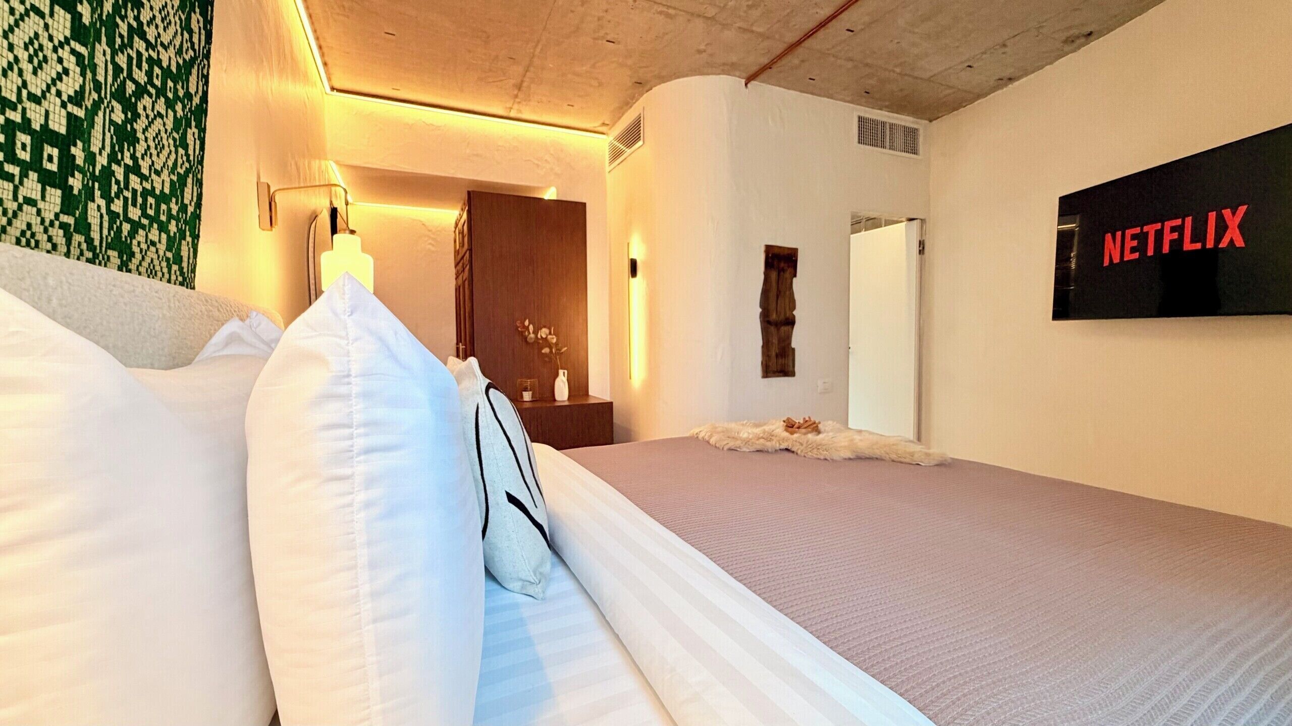 A hotel room with a bed, a wall-mounted TV displaying 'NETFLIX,' a door, and a small table with a vase.
