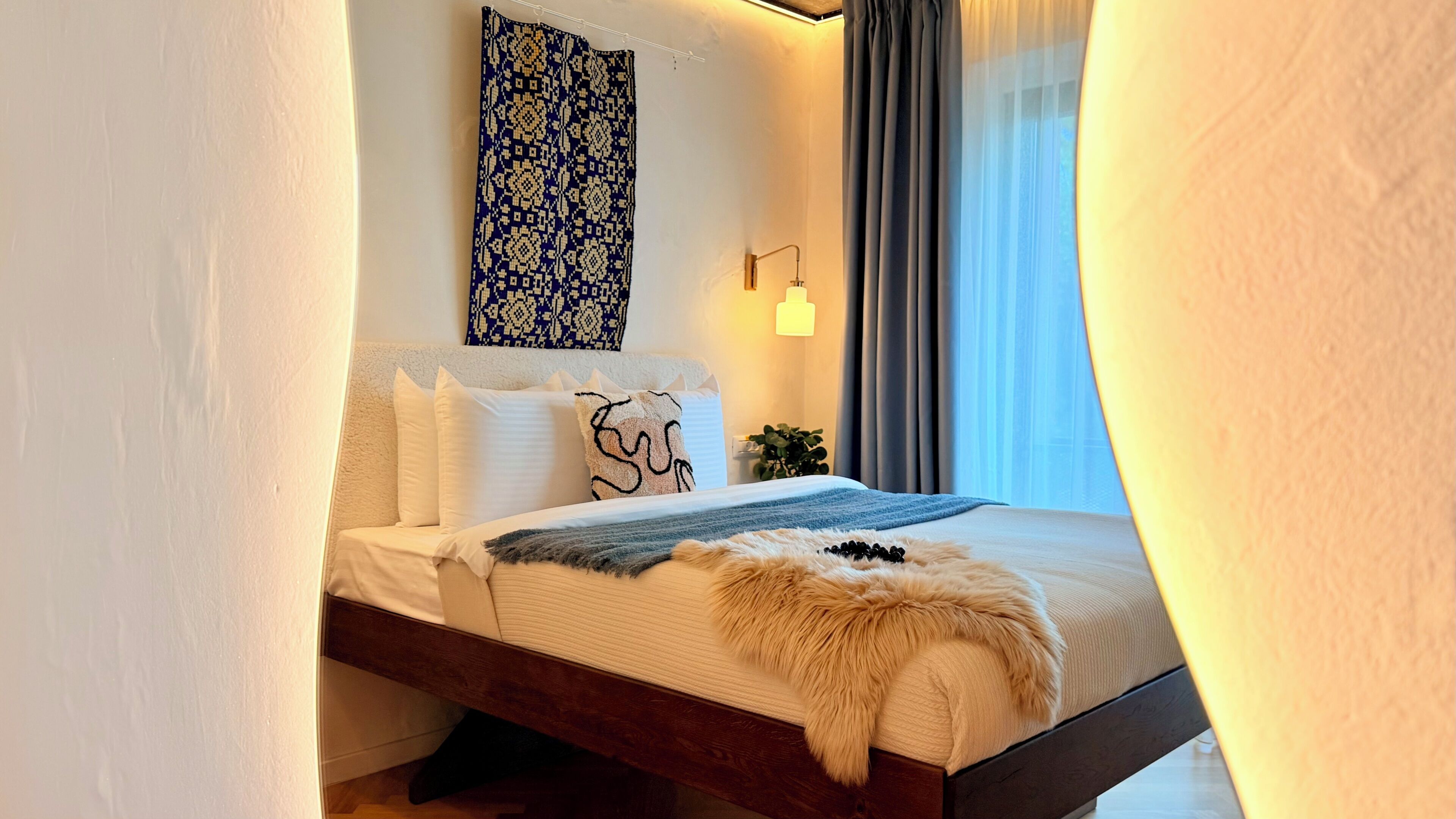 Une chambre à coucher avec un lit, des oreillers, une couverture, une lampe fixée au mur et une fenêtre avec des rideaux.