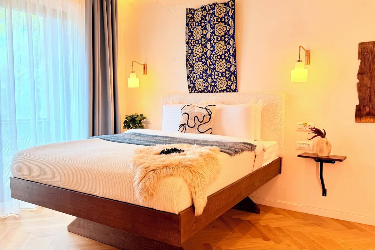A modern bedroom with a bed, bedside lamps, a wall-mounted light fixture, a decorative wall piece, and a small plant on a nightstand.