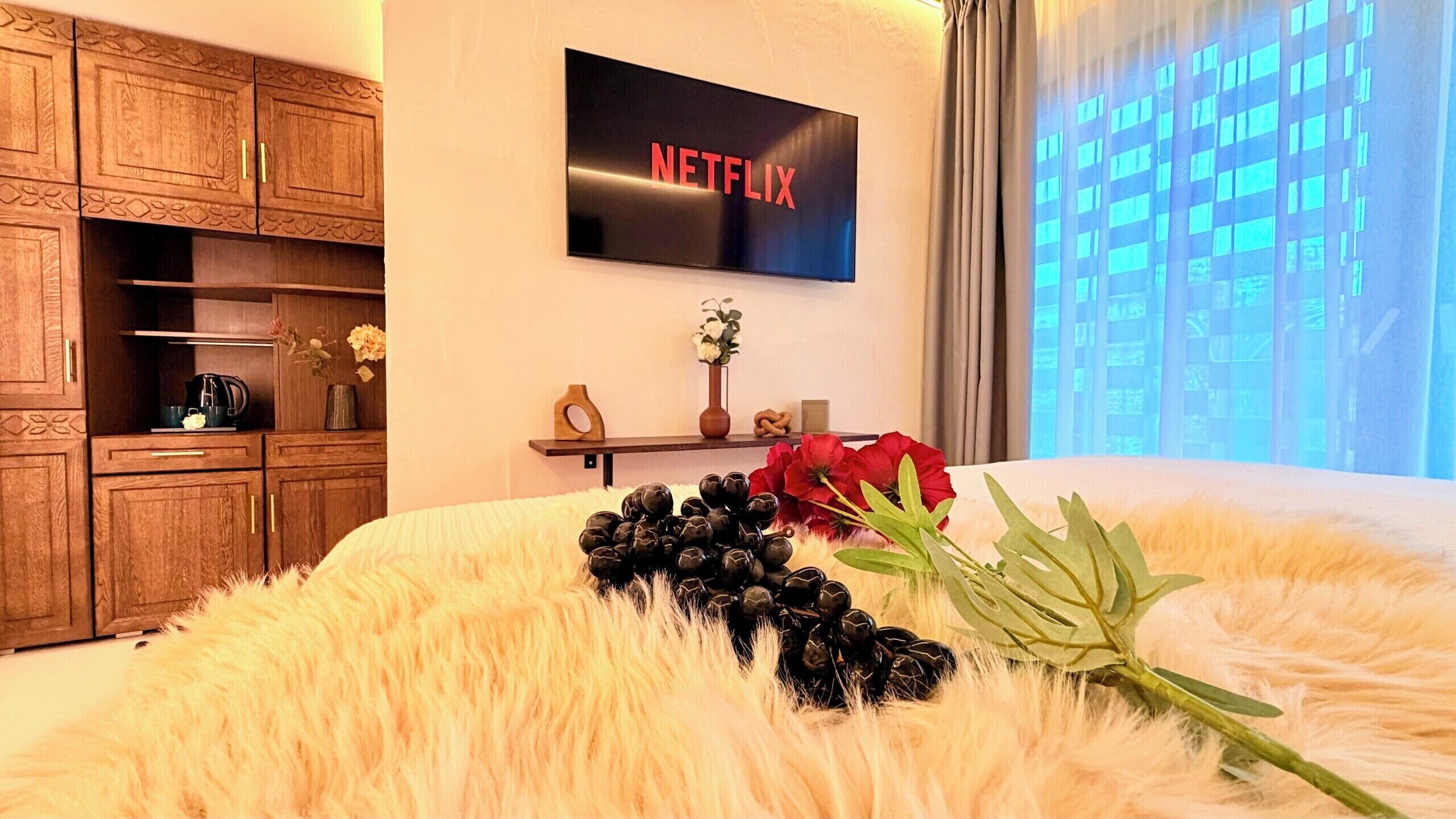 A bedroom with a bed, a TV mounted on the wall, wooden cabinets, and a window with a view.