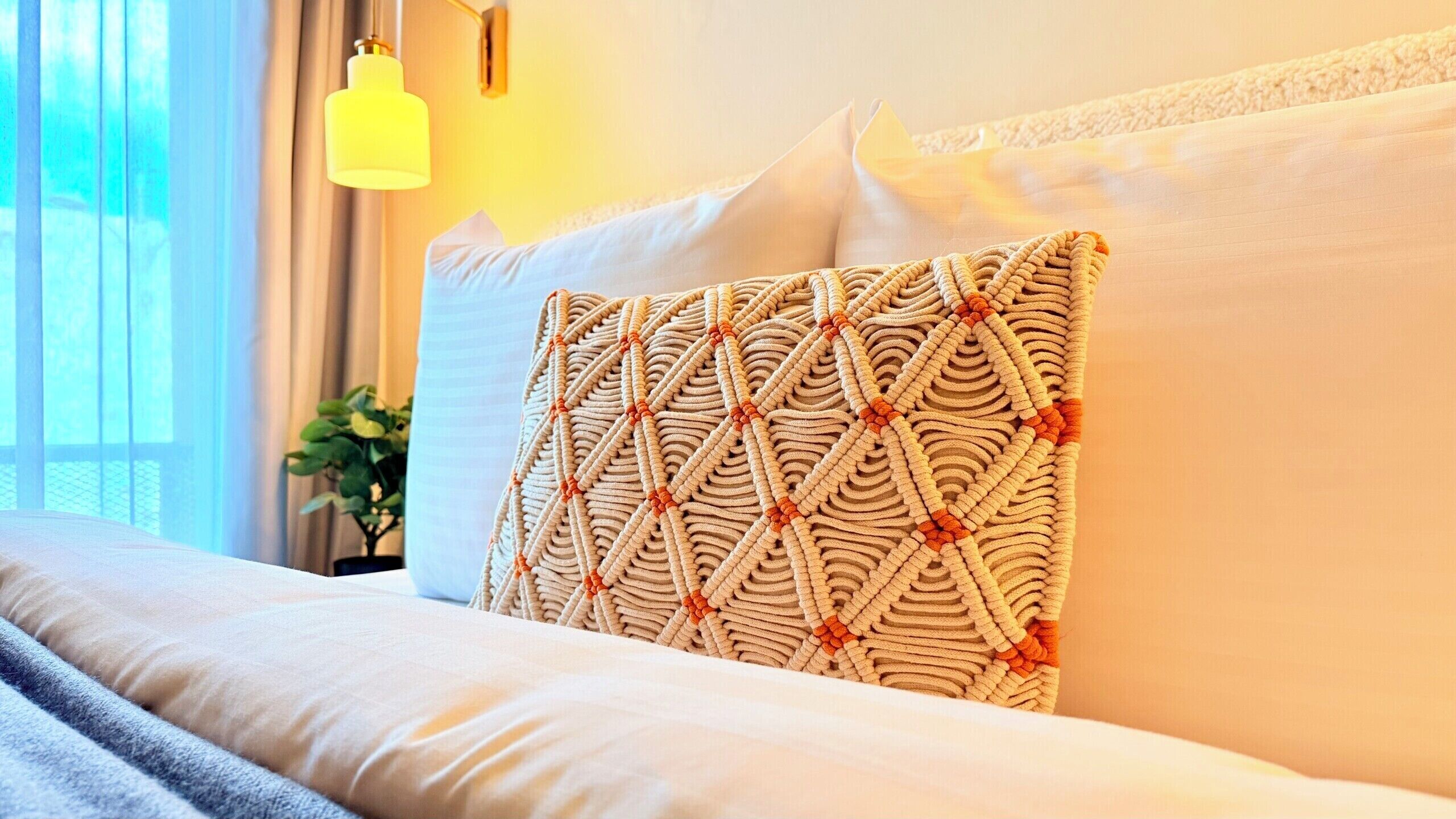 Un lit bien fait, agrémenté d’un coussin à motifs, d’une lampe de chevet et d’une fenêtre avec des rideaux.