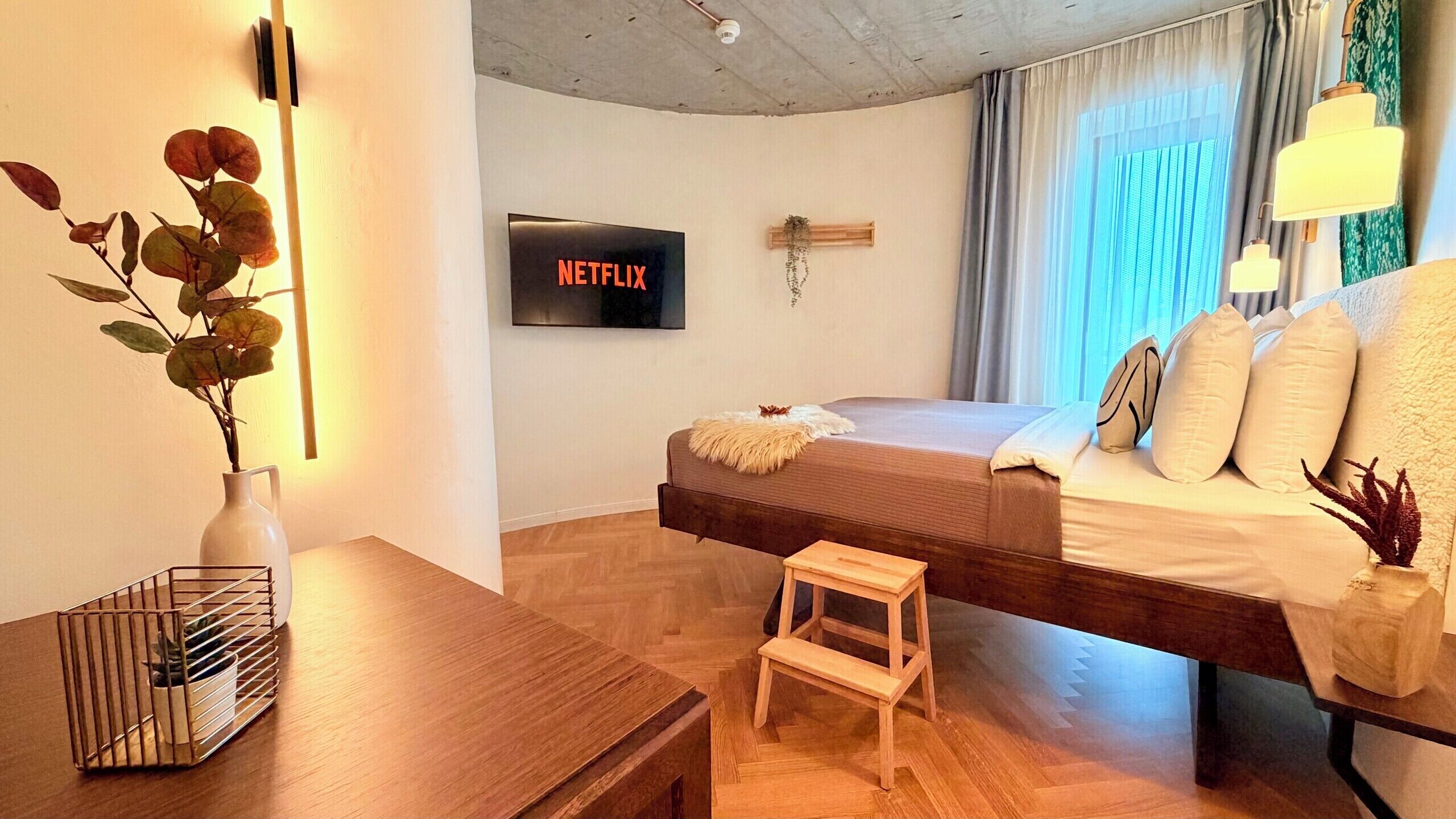 A modern bedroom with a bed, a small wooden stool, a wall-mounted TV, and a plant.