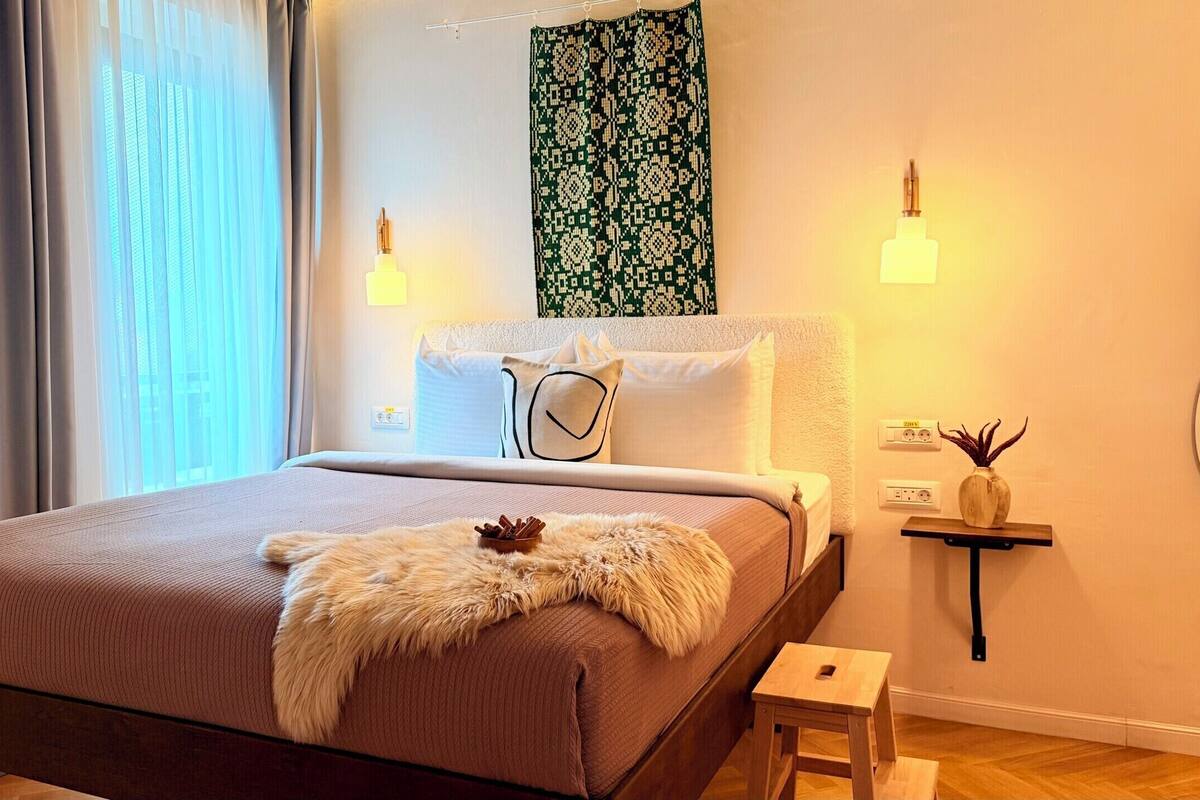 A modern bedroom with a bed, a small wooden stool, a wall-mounted plant, and a patterned tapestry.
