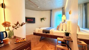 Une chambre d’hôtel moderne équipée d’un lit, d’un bureau, d’une chaise, d’un téléviseur affichant « NETFLIX » et d’une fenêtre avec des rideaux.