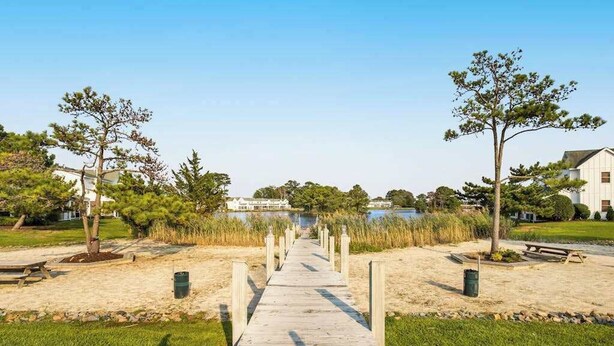 A wooden boardwalk leading to a body of water, flanked by trees and grassy areas.