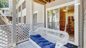 Une chaise suspendue en osier blanc se trouve sur une terrasse en bois, agrémentée d’une clôture blanche à treillis. Une porte-fenêtre coulissante donne sur une cuisine, et le plafond est en bois.