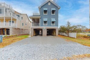 A two-story building with a garage entrance, a balcony on the upper floor, and a gravel driveway. - Downtown Bethany Beach house w/ gas grill, outside shower and screened porch (Bethany Beach)