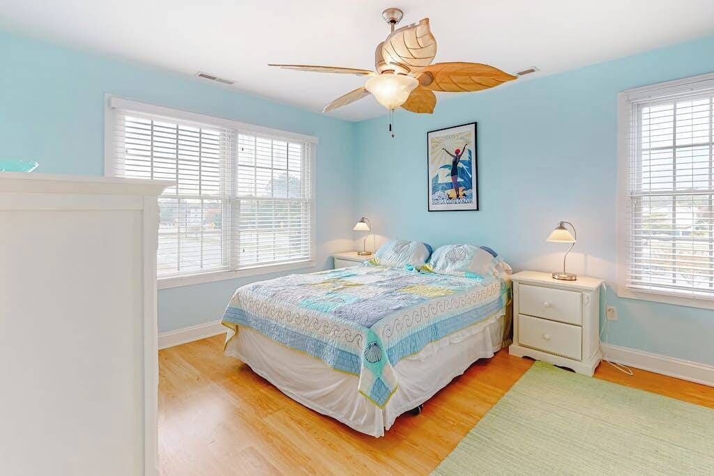 Ein Schlafzimmer mit einem Bett, zwei Nachttischen, einem Deckenventilator und zwei Fenstern mit Jalousien.