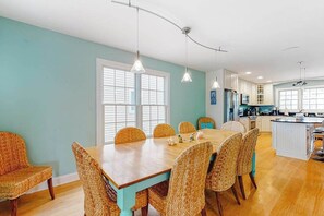 A dining area with a wooden table and wicker chairs, a kitchen with white cabinets, and a large window providing natural light. - Downtown Bethany Beach house w/ gas grill, outside shower and screened porch (Bethany Beach)