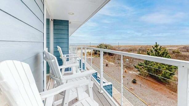 Ein Balkon mit weißen Adirondack-Stühlen, einem blauen Geländer und Blick auf Strand und Dünen.