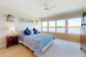 Ein Schlafzimmer mit Bett, Nachttischen, einem Schminktisch und einem Deckenventilator, mit großen Fenstern und Blick auf ein Gewässer.