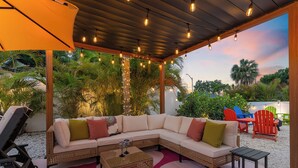 A covered outdoor seating area with a sofa, chairs, and a table, surrounded by greenery and string lights. - Hippie House-5 bedroom, Sleeps 12! Pool, Basketball, Mini golf, Pickleball Ct! (Bradenton)