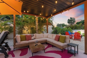 A covered outdoor seating area with a sofa, chairs, and a table, surrounded by greenery and string lights. - Unique and FUN 5-bedroom house with Pool, Basketball, Mini golf, Pickleball Ct! (Bradenton)