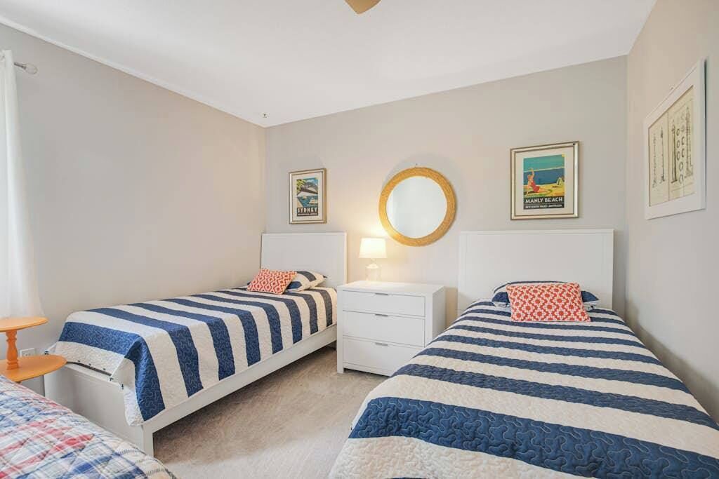 A bedroom with two single beds, a bedside table with a lamp, a round mirror, and two framed pictures on the wall.
