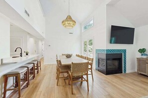 A spacious dining area with a long table and chairs, a kitchen with a white countertop, and a fireplace with a tiled surround. - Corner Sea Colony condo with screened porch, pools, hot tub & tennis (Bethany Beach)