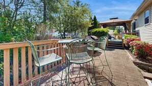 Eine Terrasse mit Metallstühlen und -tischen, ein Holzboden und ein überdachter Bereich mit Treppen, die zu einem Haus führen.