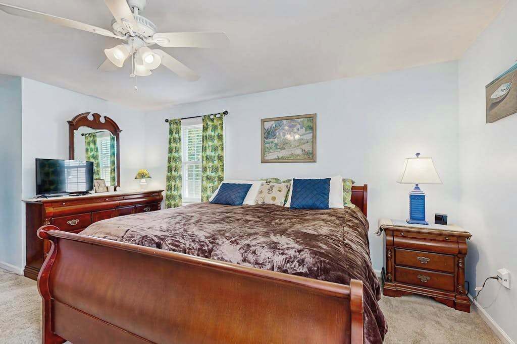 A bedroom with a bed, a ceiling fan, a dresser, a nightstand, a lamp, a TV, and a window with curtains.