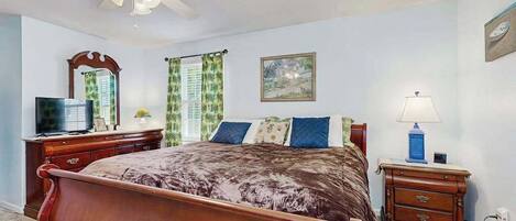 A bedroom with a bed, a ceiling fan, a dresser, a nightstand, a lamp, a TV, and a window with curtains.