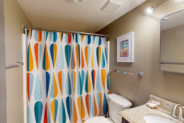 Casa de banho com cortina de chuveiro com padrão abstrato colorido, sanita, lavatório com bancada de granito e um quadro na parede.