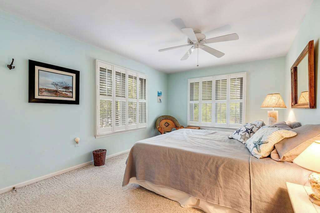 Ein Schlafzimmer mit einem Bett, einem Deckenventilator, zwei Fenstern mit Rollläden, einem gerahmten Bild und einer Lampe.
