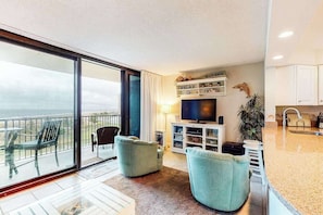 A modern living room with a TV, seating, and a balcony with a view. - Sea Colony 3rd floor condo w/ elevator, shared tennis court & gym - ocean view (Bethany Beach)