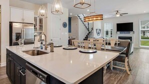 A modern kitchen with a large island, stainless steel appliances, pendant lighting, and a dining area with chairs and a table. - Ocean View Beach Club stylish home with outdoor pool, fast WiFi, & wet bar (Ocean View)