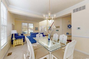 A dining area with a glass table and white chairs, a chandelier, and a blue sofa in the background. - Bethany Lakes house w/ private gas grill, balcony, free WiFi, and pool (Bethany Beach)