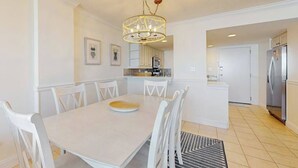 Un comedor con una mesa y sillas blancas, una araña y una cocina al fondo.