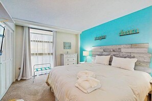 A hotel room with a bed, a nightstand, a lamp, a window with curtains, a wall with a 'dream' sign, and a small table with a chair. - Sea Colony oceanfront 2nd floor condo w/ free WiFi, shared sauna, and pool (Bethany Beach)