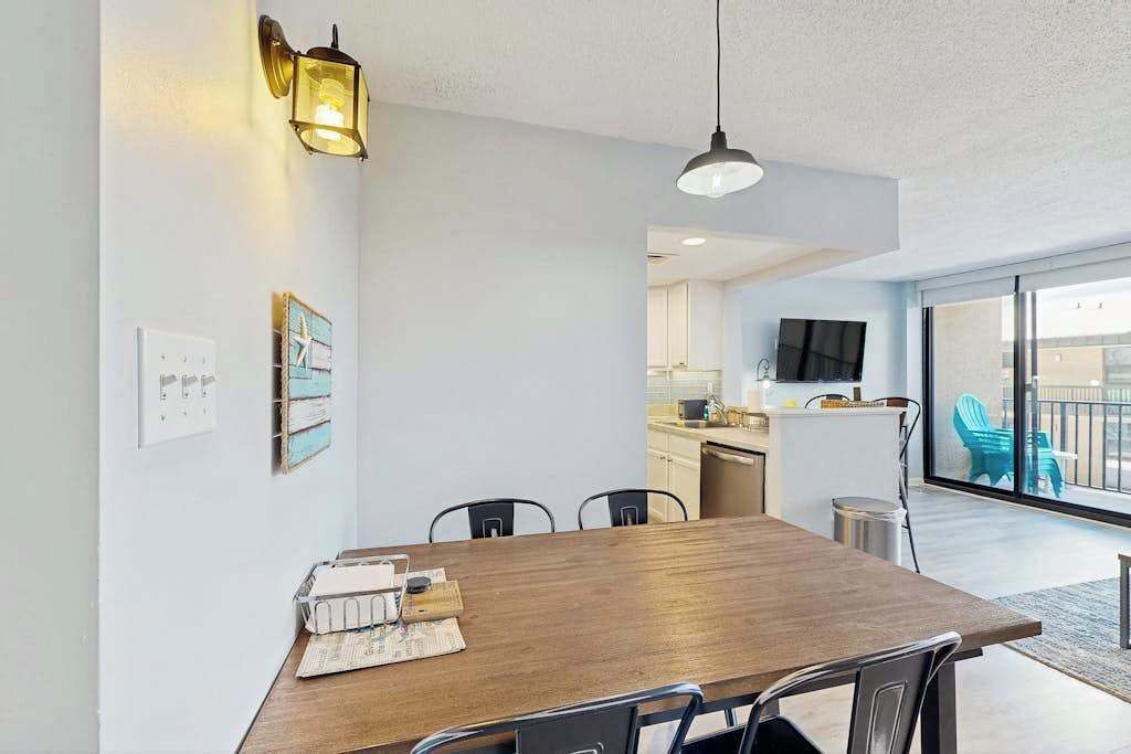 A modern dining area with a wooden table, chairs, a pendant light, and a view of the kitchen and balcony.