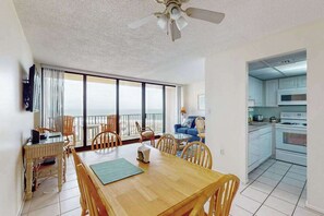 Ein Zimmer mit Esstisch, Stühlen, Deckenventilator, einer Küche mit weißen Schränken und Blick durch große Fenster.