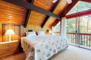 Ein Schlafzimmer mit Holzboden, einem Bett mit floralem Überwurf, einem Nachttisch mit Lampe und einem großen Fenster mit Blick auf Bäume.