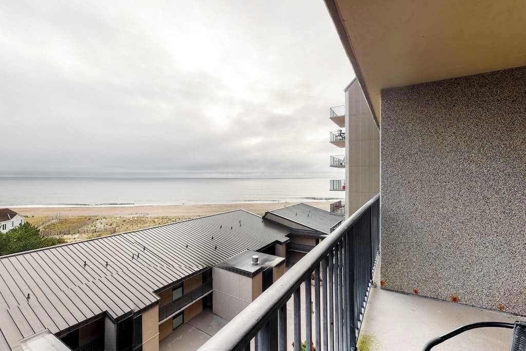 Een balkon met uitzicht op het strand en de oceaan, een gebouw met een grijze gevel en een metalen dak.