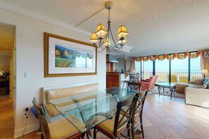 A spacious living area with a glass dining table, a sofa, and a balcony with a view. - Sea Colony 1st floor condo w/ pool, gym, and balcony - ocean view (Bethany Beach)