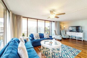Ein Wohnzimmer mit Sofa, Sesseln, Couchtisch, Fernseher und einem Balkon mit Vorhängen.