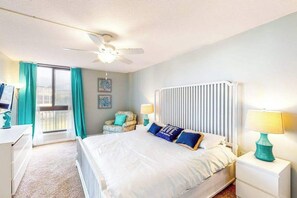 Une chambre d’hôtel comprenant un lit, un bureau, une chaise, une fenêtre avec des rideaux, un radiateur et un ventilateur de plafond.