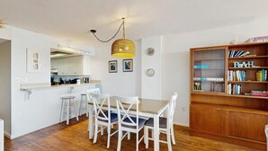 A compact kitchen with white cabinets, a dining table with white chairs, a hanging light fixture, and a bookshelf filled with books. - Sea Colony Oceanside Condo Steps to Private Beach, Pools and Tennis (Bethany Beach)
