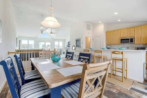 A spacious dining area with a wooden table and blue-striped chairs, a kitchen with wooden cabinets, and a view of the outside through large windows. - Villas at Beach Cove Getaway W/ Bay/Ocean Views, Private Balcony & Shared Pool (Bethany Beach)