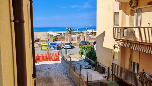 Une vue depuis un balcon donnant sur une rue pavée, avec un ciel bleu dégagé et une plage qui s’étend au loin.
