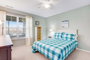 Une chambre à coucher comprenant un lit, un bureau, un ventilateur de plafond, une fenêtre avec des stores et un panneau décoratif mural.
