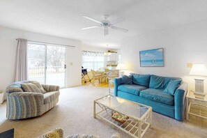 A living room with a blue sofa, a patterned armchair, a glass coffee table, a ceiling fan, and a view of the kitchen and outdoor area. - Clear Water Home Close to the Beach w/Shared Pool, Private Washer/Dryer (Frankford)