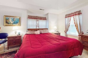 Un dormitorio con una cama grande, dos mesitas de noche con lámparas, una silla, un aparador, un cuadro en la pared y cortinas en las ventanas.