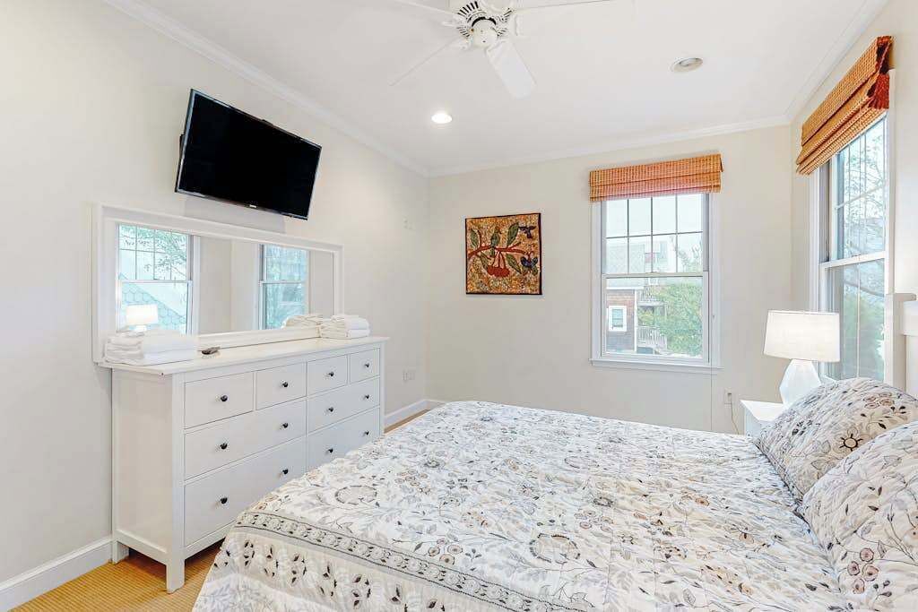 Un dormitorio con cama, cómoda, televisor montado en la pared y ventana con vistas.