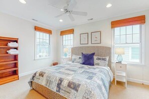 A bedroom with a bed, a ceiling fan, a nightstand, a lamp, a wooden shelf, and a window with a view. - Town of Bethany beach house with expansive rooftop deck & several kitchens (Bethany Beach)