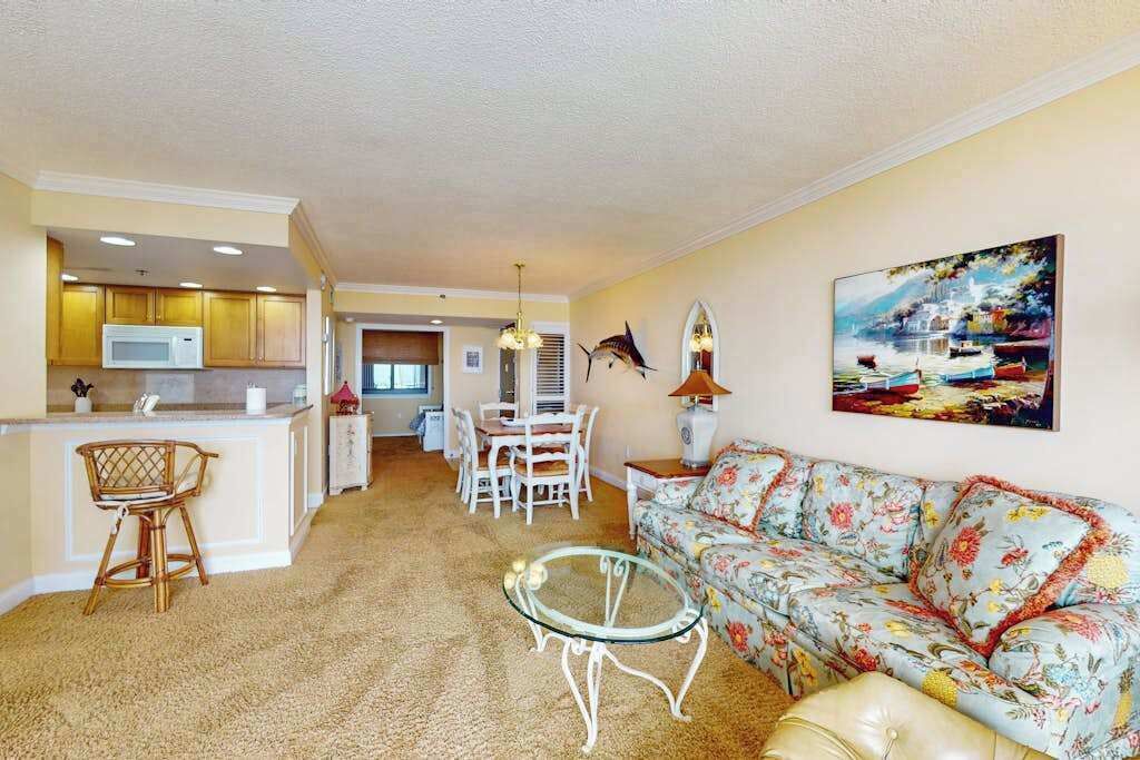 A living room with a sofa, a glass coffee table, a dining table with chairs, a kitchen area with a microwave, and a painting on the wall.